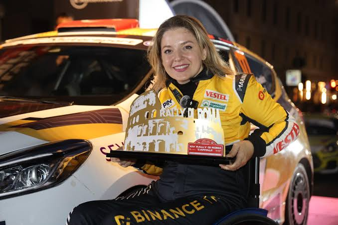 Smiling woman in a yellow 'VESTEL' racing suit holding a 'Rally of Roma Capitale' trophy in front of a rally car.