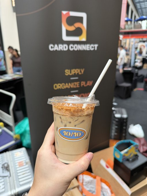 A hand holds a cup of iced coffee with a "10/10" sticker in front of a "Card Connect" booth at an event, where the banner displays the text "SUPPLY ORGANIZE PLAY."