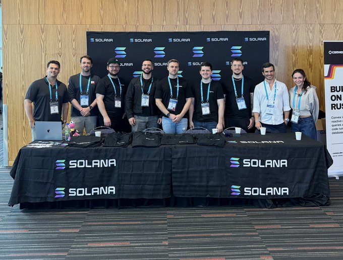 A group of nine people posing behind a table draped with black 'SOLANA' branded tablecloths, standing in front of a matching Solana backdrop at an event.