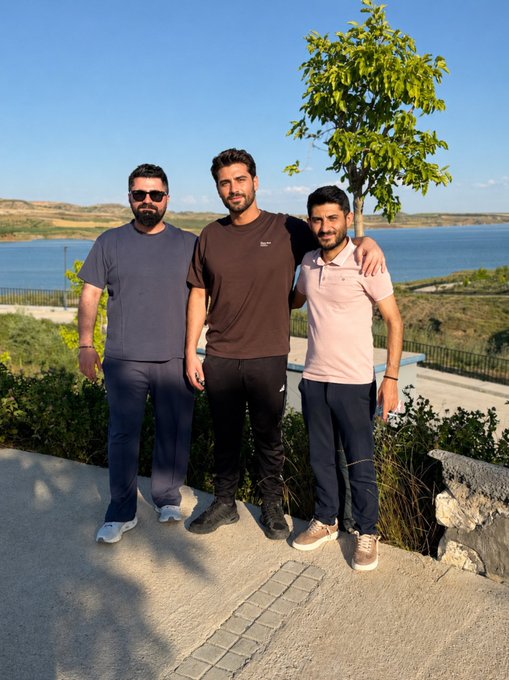 The image shows three men standing together, posing for a photo near a body of water. The man on the left has his arm around another man who is wearing a pink shirt. The third man stands next to them with his arm around the second man's shoulder. They are all smiling and enjoying their time together in front of the lake.