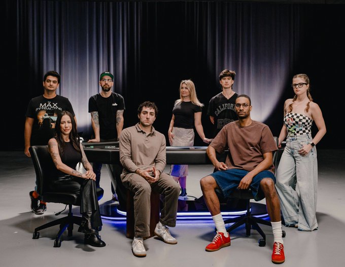 A group portrait of eight people posing in a dark studio with curtains and a glowing blue-lit table, dressed in casual streetwear including one man in a "MAX" shirt, another in "California" apparel, and a woman in a checkered top.