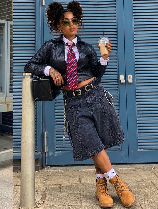 A stylish woman poses outdoors against a blue shuttered wall, wearing a black leather jacket, striped tie, denim skirt, sunglasses, and holding an iced coffee.
