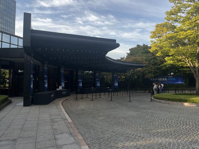 An outdoor venue entrance features a modern black architectural canopy with banners and signage reading 'K-POP SEOUL 2020' on a paved area.