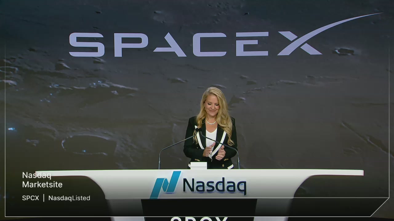 The image features a woman standing behind a podium, giving a speech or presentation at an event. She appears to be wearing a suit and has blonde hair. The background of the image shows a large screen displaying the words "SPACEX" and "Nasdaq", indicating that this is likely a conference or event related to space exploration or stock market activities.