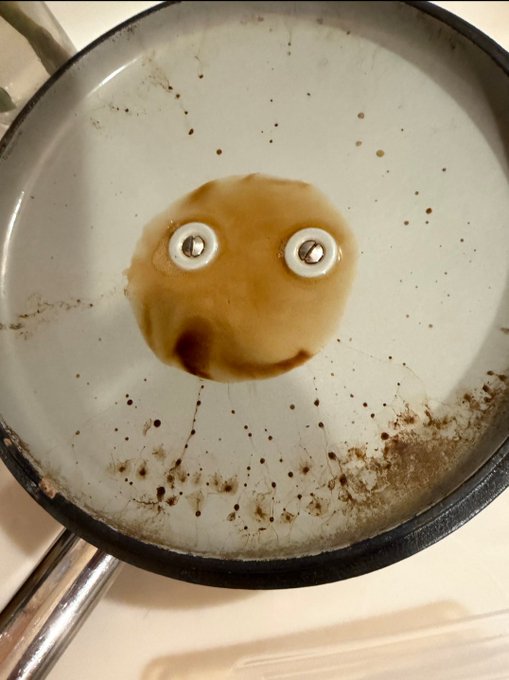 A dirty white frying pan containing a small puddle of brown liquid that looks like a face, created with two googly eyes placed on the pan.