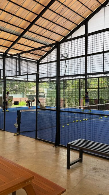 An indoor padel/tennis court with blue flooring where two people are playing, with a bench and table visible in the foreground.