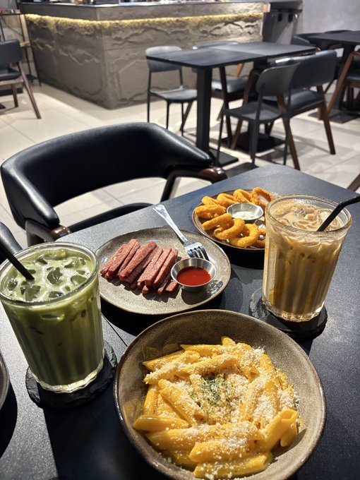 A table is set with matcha latte, iced coffee, french fries, penne pasta, sliced meat, and onion rings in a restaurant.