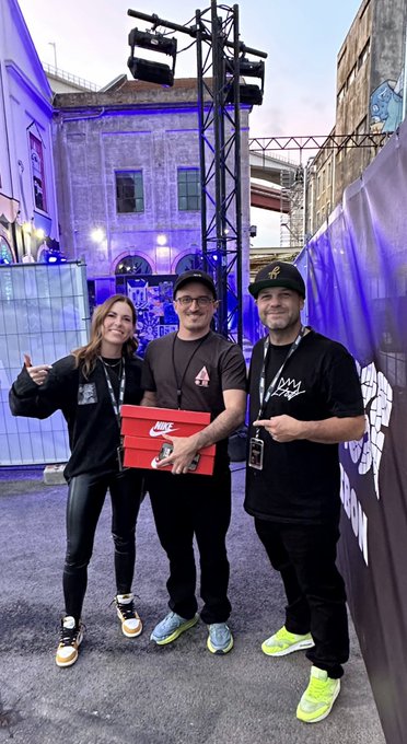 Three people pose outdoors, with the man in the middle holding a red Nike shoebox.