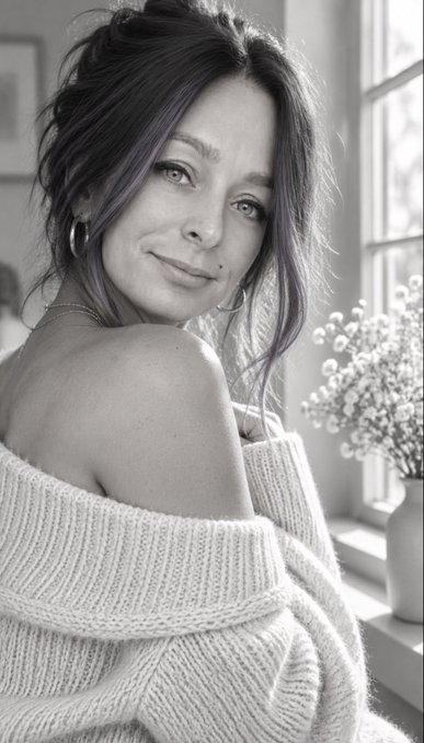 A black-and-white portrait of a woman with long wavy hair wearing an off-shoulder knit sweater, posed by a window with baby's breath flowers.