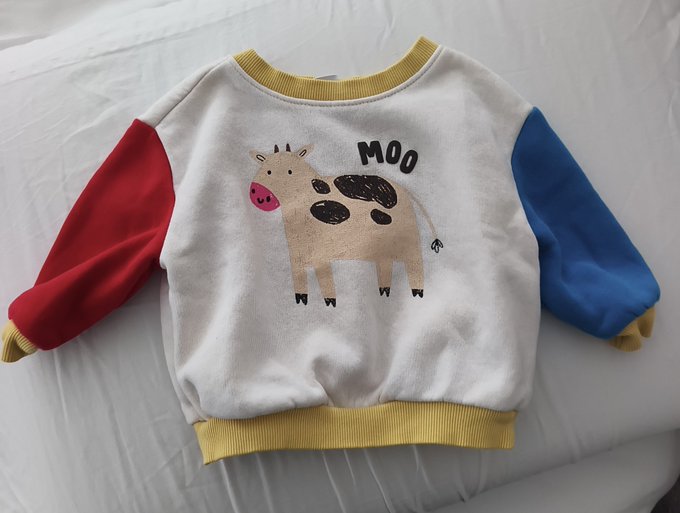 A children's sweatshirt laid flat on a white sheet features a cream body with red and blue sleeves, yellow trim, and a cartoon cow design with the word 'MOO'.