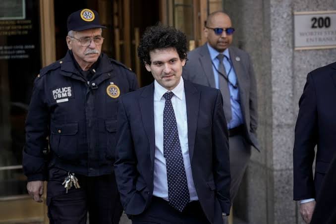 Sam Bankman-Fried, wearing a dark suit and patterned tie, exits 200 Worth Street (sign visible in the background) flanked by a U.S. Marshals Service officer and security personnel, in an image that relates to the post about his fraud conviction appeal and ongoing legal battles.