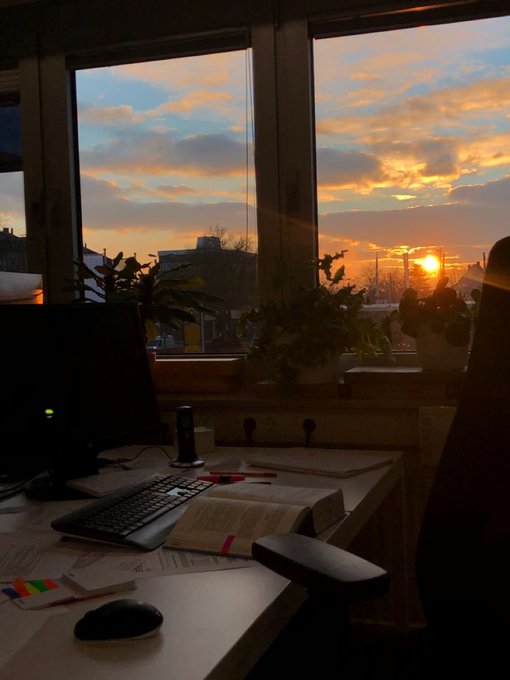The image shows an office desk with various items, including a computer monitor, keyboard, mouse, and a book or notebook. The setting appears to be a workspace where someone might work from home during the day. A window can also be seen in the background, providing natural light for the room. The photo is taken at sunset, creating a serene atmosphere with warm hues of orange and yellow lighting.