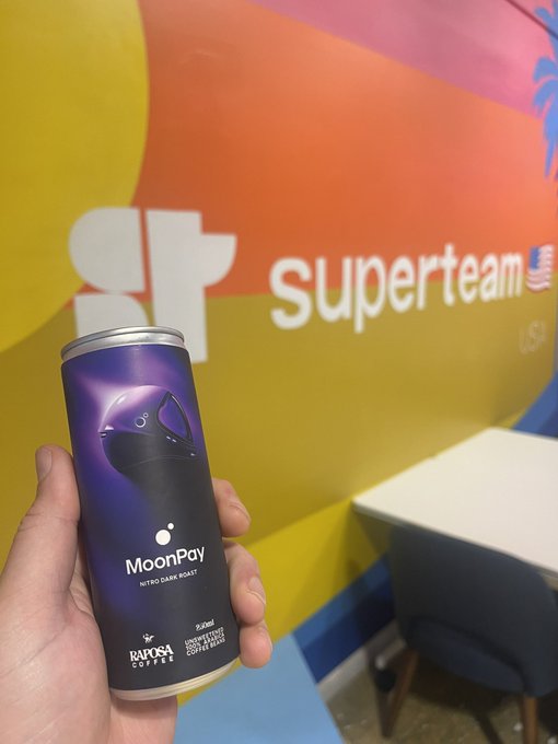 A hand holds a can of MoonPay Nitro Dark Roast coffee by Raposa Coffee, photographed in front of a colorful "superteam US" branded wall.