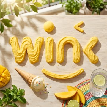 The image displays yellow frosting or batter artfully shaped into the word "wink" on a wooden surface, surrounded by tropical accents like a mango, ice cream cone, and lime drink. The word "wink" appears to be a playful nod to the post about Wind-powered water pumping, where "Win" cleverly ties into the "wind" theme of the community irrigation grants on TRON blockchain.