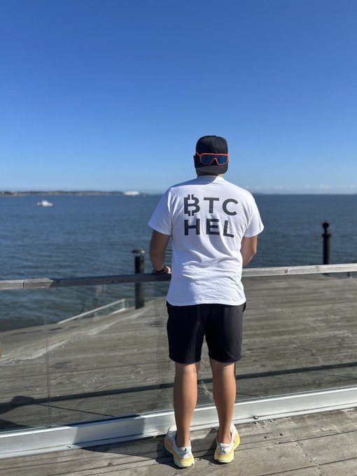 Photo of a man from behind wearing a white t-shirt with 'BTC HEL' (Bitcoin logo) text, black shorts, ski goggles, and yellow sneakers, standing on a wooden pier looking at the ocean.