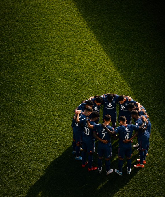 A soccer team in navy blue "crypto.com" jerseys huddles together on a sunlit green field, casting long shadows across the pitch. The image reflects the post's message of unity and strategic focus, capturing a moment of teamwork before the players take their positions and make their next move on the field.