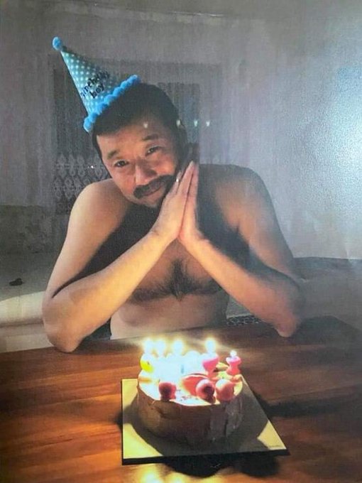 A shirtless man wearing a blue 'Happy Birthday' party hat sits with his hands clasped in front of a lit birthday cake, appearing to be in a dimly lit room.