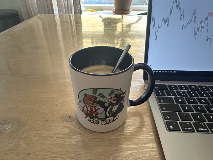 A 'I LOVE TRADING' cartoon mug with a spoon sitting on a wooden desk next to a laptop displaying a line chart.