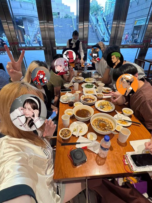 A group of people enjoy a meal at a large restaurant table filled with various dishes, their faces covered with circular anime-style stickers; one person flashes a peace sign.