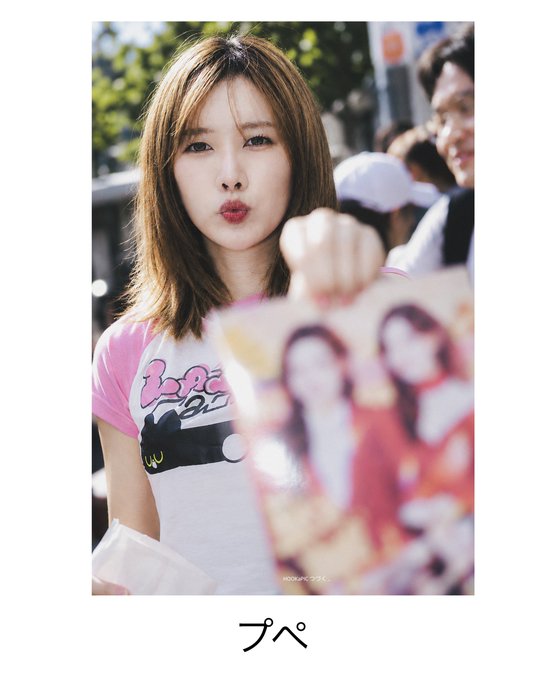 A young woman with shoulder-length brown hair poses with a pouty expression, holding up a pink photo card or album featuring illustrated characters; the image is captioned "プペ" (Pupe) at the bottom.