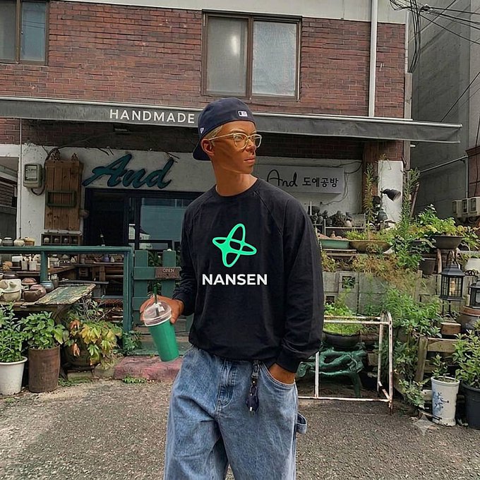 A young man wearing a blue cap, yellow glasses, and a black sweatshirt with a green atom logo and the text 'NANSEN' stands in front of a brick building with 'HANDMADE' and 'And' signage.