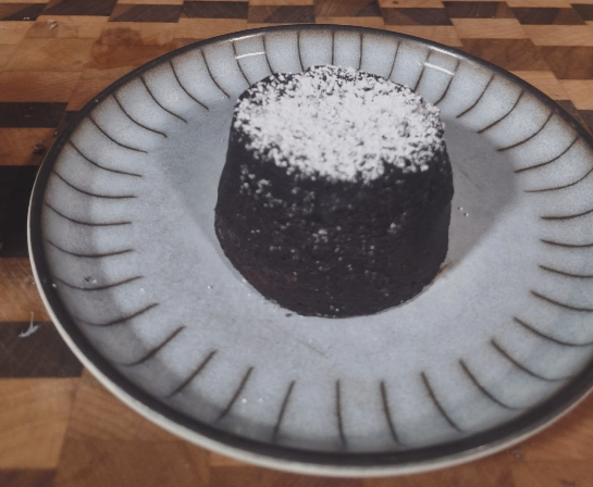 A dark cylindrical dessert dusted with powdered sugar sits on a gray ceramic plate on a wooden table.