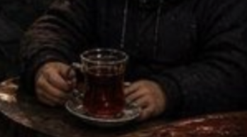 A dimly lit scene showing a person in a dark jacket seated at a table, holding a traditional Turkish tea glass in a tulip-shaped holder on a saucer.