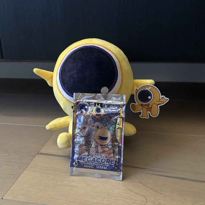 A yellow plush toy with a large purple eye and a small matching sticker sit on a wooden floor in front of a trading card pack labeled 'WINK'S MEGACORE MMH' featuring cartoon monster illustrations.