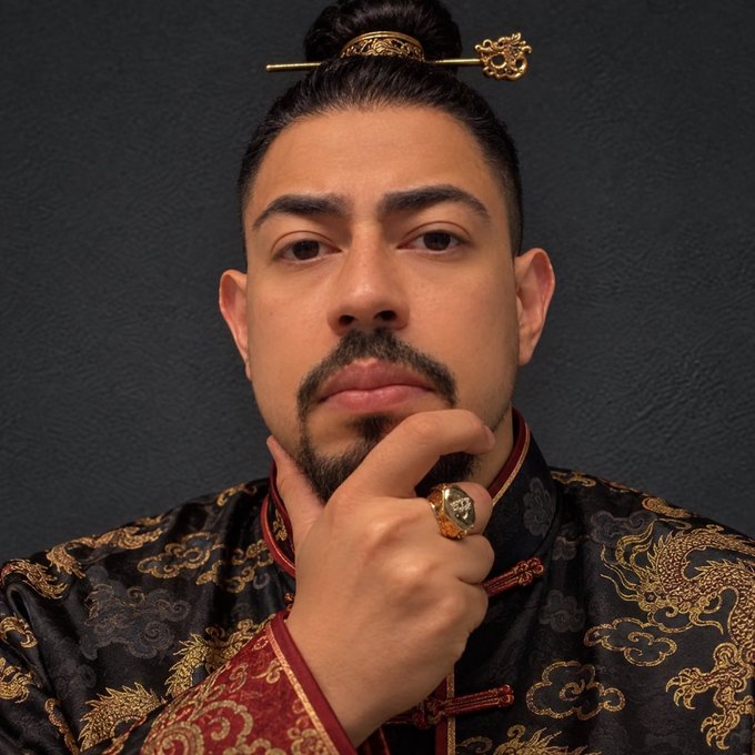 A man wearing a traditional Chinese silk robe with gold dragon embroidery and a decorative hairpin poses thoughtfully with his hand on his chin, embodying the "Founder from China" persona referenced in the caption while appearing contemplative about his next memecoin venture.
