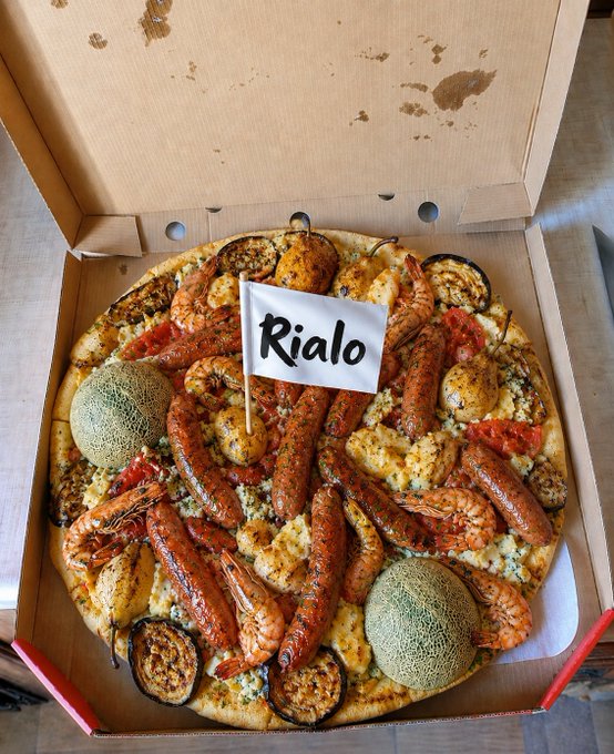 A gourmet pizza in a cardboard box topped with shrimp, sausage, and other ingredients, featuring a small white flag in the center with the name 'Rialo' written on it.