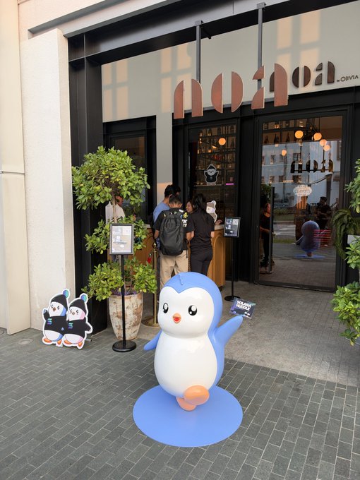 Outside a 'mother' storefront by Obiya, a blue and white penguin statue stands on a blue platform beside a smaller cutout, with people entering the shop in the background.