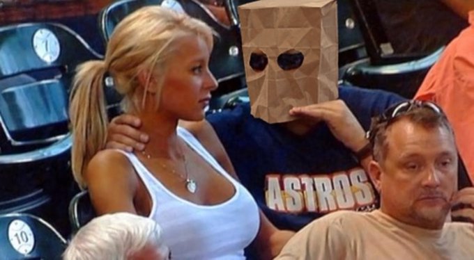 The image shows a man wearing a geometric cardboard "baghead" mask sitting in stadium seats next to a blonde woman in a white top, with another man beside him wearing an "ASTROS" shirt—suggesting a Houston Astros baseball game. The viral "bag guy" meme humorously parallels the post's theme of a quirky, anonymous crypto/token project where users are rewarded for holding while the token burns supply to drive value up, mirroring the absurdity of the masked man's unusual appearance at a casual sporting event.