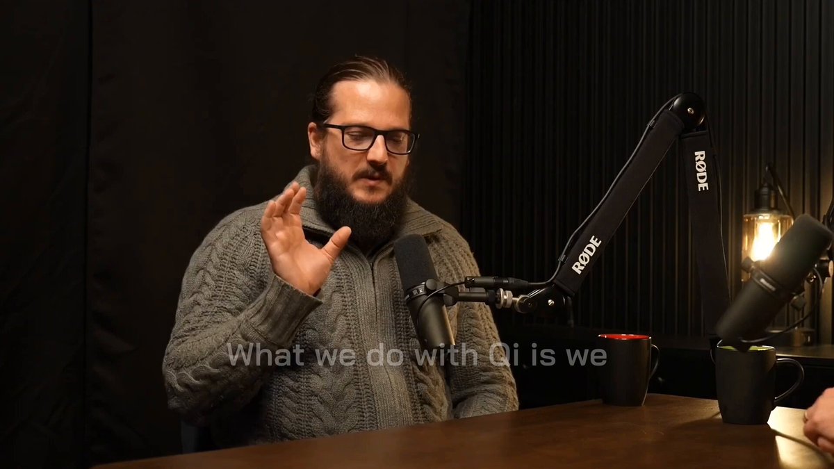 The image features a man wearing glasses sitting at a table with a microphone, likely participating in an interview or discussion. He has his hand raised above his head while talking into the microphone. The background of the image shows a black curtain, which could be part of a recording studio setup. There are two cups placed on the table next to him and another cup further away from him. The text "Dr. K explains private $QI transactions" is visible in the image, possibly indicating that Dr. K is discussing private transactions or QI (Quality International) with someone else who is not present in the frame.