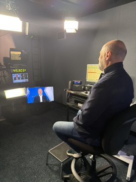 A bearded man in a dark jacket sits in a broadcast control room filled with monitors and studio lighting, with one screen displaying "WPBS" graphics and another showing a person in a blue suit, suggesting he is preparing for an upcoming program or announcement teased by the "Coming soon." caption.