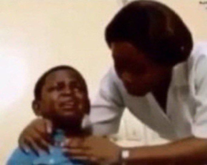 A blurry photo showing two people, with a young boy in a blue shirt sitting while a woman in a white top appears to be touching or examining his face.