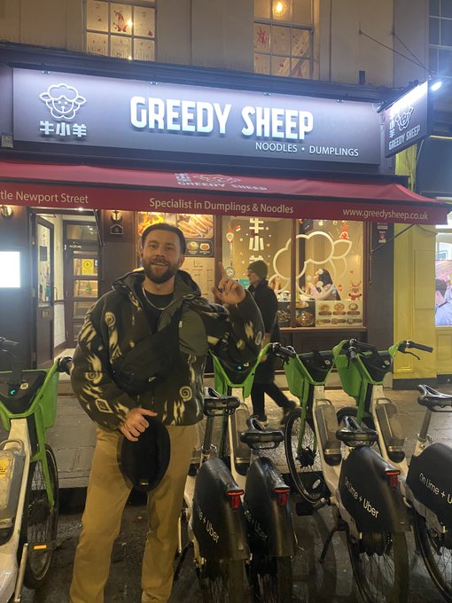 A man wearing a camouflage hoodie and tan pants stands on a street at night in front of 'GREEDY SHEEP NOODLES + DUMPLINGS' restaurant with green rental bikes lined up beside him.