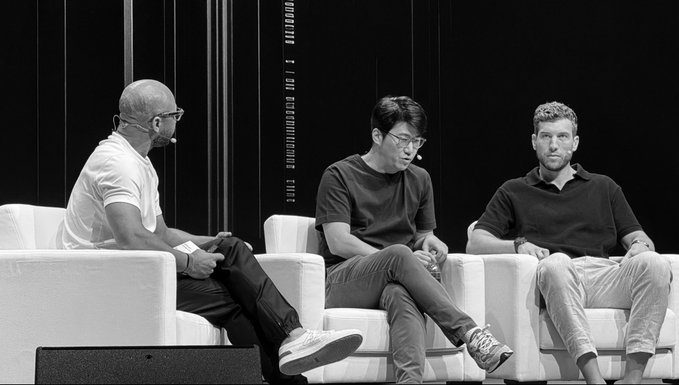 Three men seated on white chairs on a black stage, appearing to participate in a panel discussion.