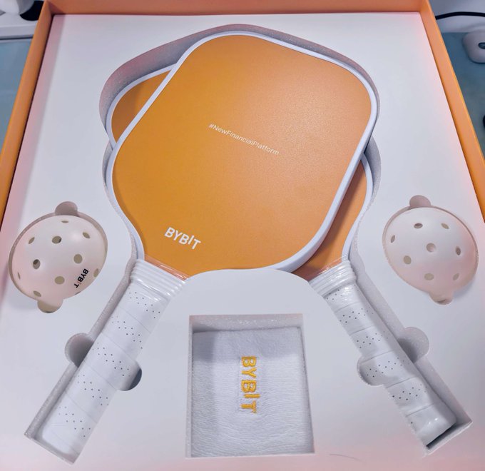A branded pickleball set in a white box featuring orange and white paddles labeled 'BYBIT' and '#NewFinancialPlatform,' accompanied by two perforated balls with 'BYBIT' branding and a small towel also marked 'BYBIT.'