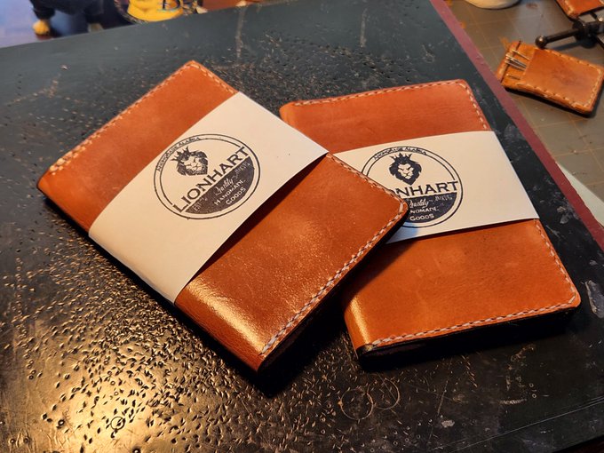 The image shows a pair of leather-bound notebooks sitting side by side, with their bindings facing up against each other. The notebooks are placed on a black surface, which contrasts with the warm tones of the leather. In the background, there is another object that appears to be a wallet or purse. The text in the image reads "Last look before shipping paid for in dogecoin Thank you for your support". This suggests that someone has recently purchased these notebooks and is about to ship them off.