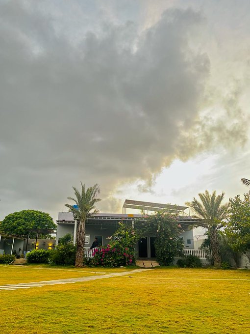 A modern white house surrounded by palm trees and blooming flowers stands under a dramatic, cloudy sky, with a lush green lawn stretching out in the foreground—perfectly capturing the "touch some grass" essence of the post's weekend message.