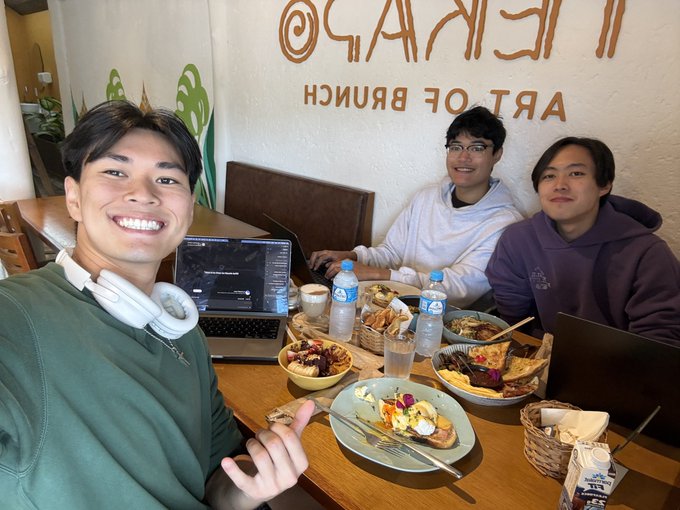 Three young men taking a selfie at an 'ART OF BRUNCH' restaurant, smiling at the camera with a laptop open and plates of colorful food on the table.