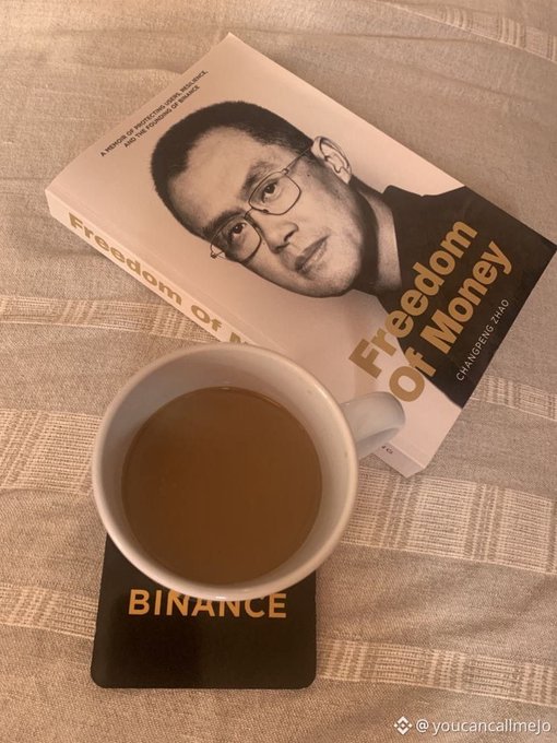 A cup of tea on a Binance coaster next to the book 'Freedom of Money' by Changpeng Zhao, subtitled 'A memoir from the man who made crypto trustworthy,' resting on a striped tablecloth.