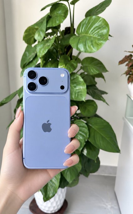 A hand with manicured nails holds a light blue iPhone with a triple-camera setup in front of a lush green potted plant.