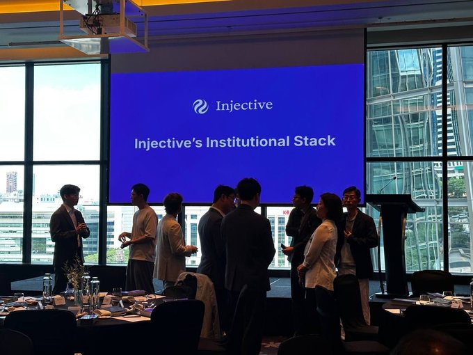 A presentation event with attendees networking in front of a large blue screen displaying 'Injective' and 'Injective's Institutional Stack.'