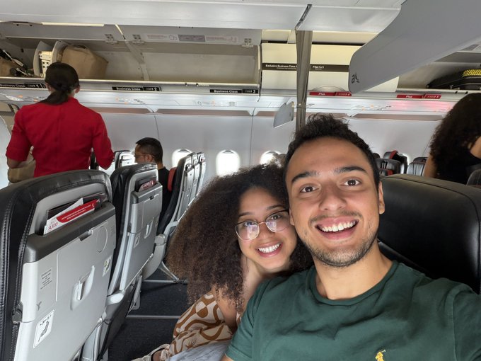 A smiling young couple taking a selfie inside an airplane cabin, with a flight attendant in red visible in the background.