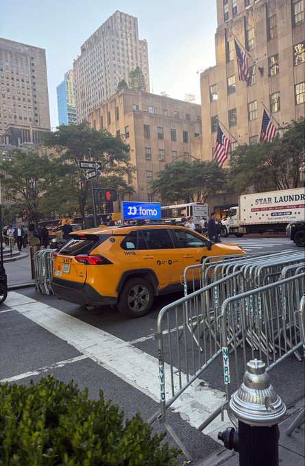 A yellow taxi parked on a New York City street near metal barricades, with American flags and tall buildings visible in the background, along with a truck labeled 'STAR LAUNDRY'.