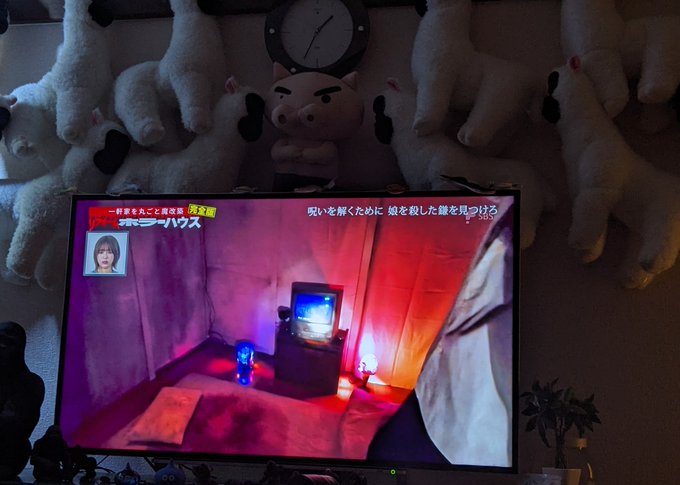 A living room scene with a TV displaying a Japanese variety show featuring a dimly lit room with red and purple lighting, surrounded by a wall of stuffed dog plushies and a clock above the television.
