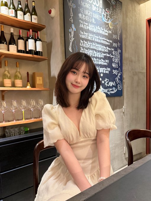 A young woman with shoulder-length brown hair wearing a cream-colored dress, seated at what appears to be a wine bar with shelves of wine bottles and a chalkboard menu visible behind her.