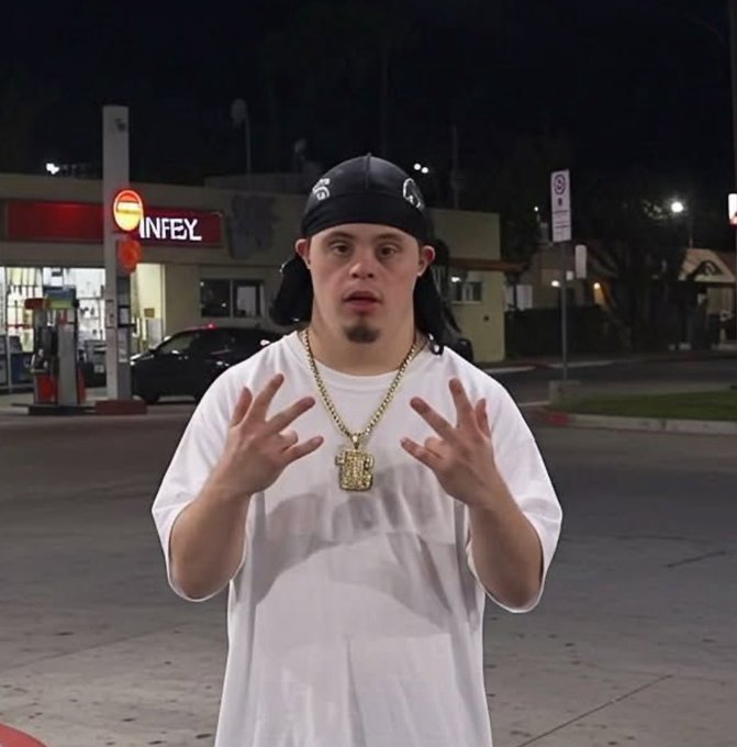 A man wearing a black bandana, white t-shirt, and gold chain pendant stands in front of a gas station at night while making hand gestures.