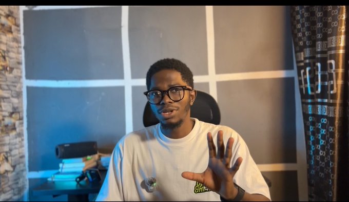 A young man wearing glasses and a white t-shirt speaks to the camera with his hand raised in a gesturing motion, seated in front of a wall with blue panels.